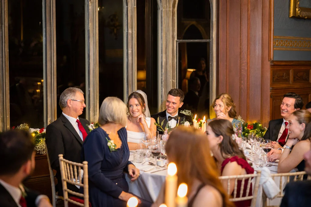 A newlywed couple sits smiling at the top table during a candlelit wedding breakfast in a historic stately home with wood-panelled walls.