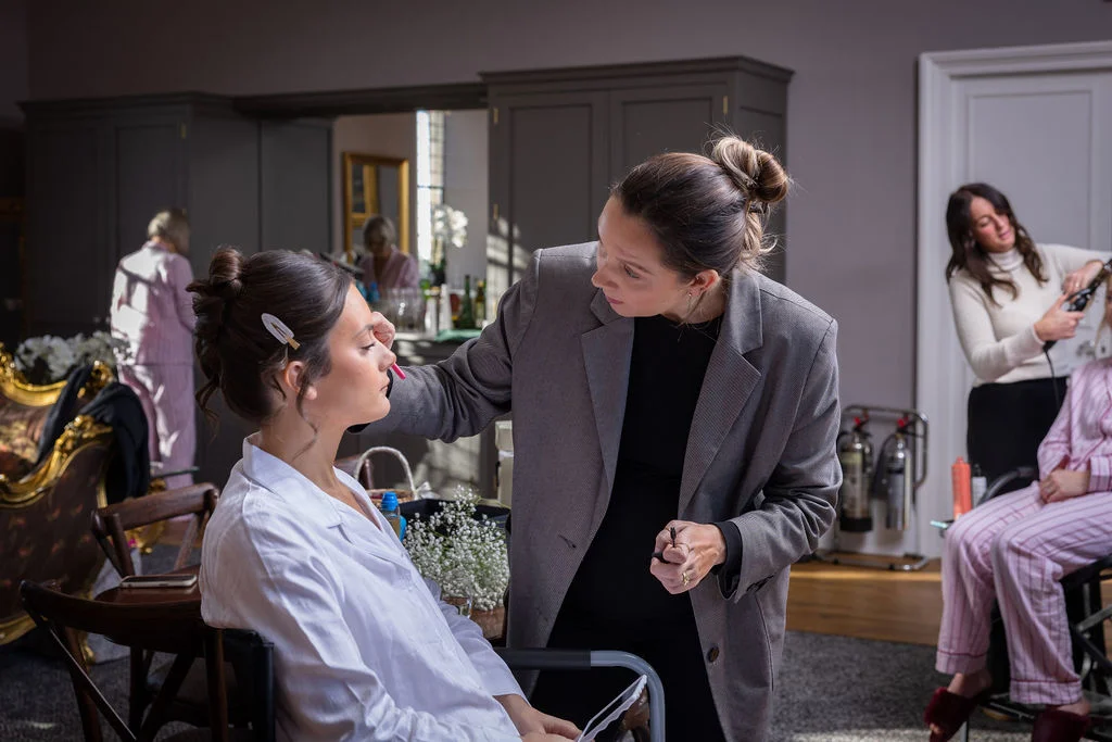A professional makeup artist applies finishing touches to a bride wearing a white robe during wedding morning preparations inside a well-lit room.