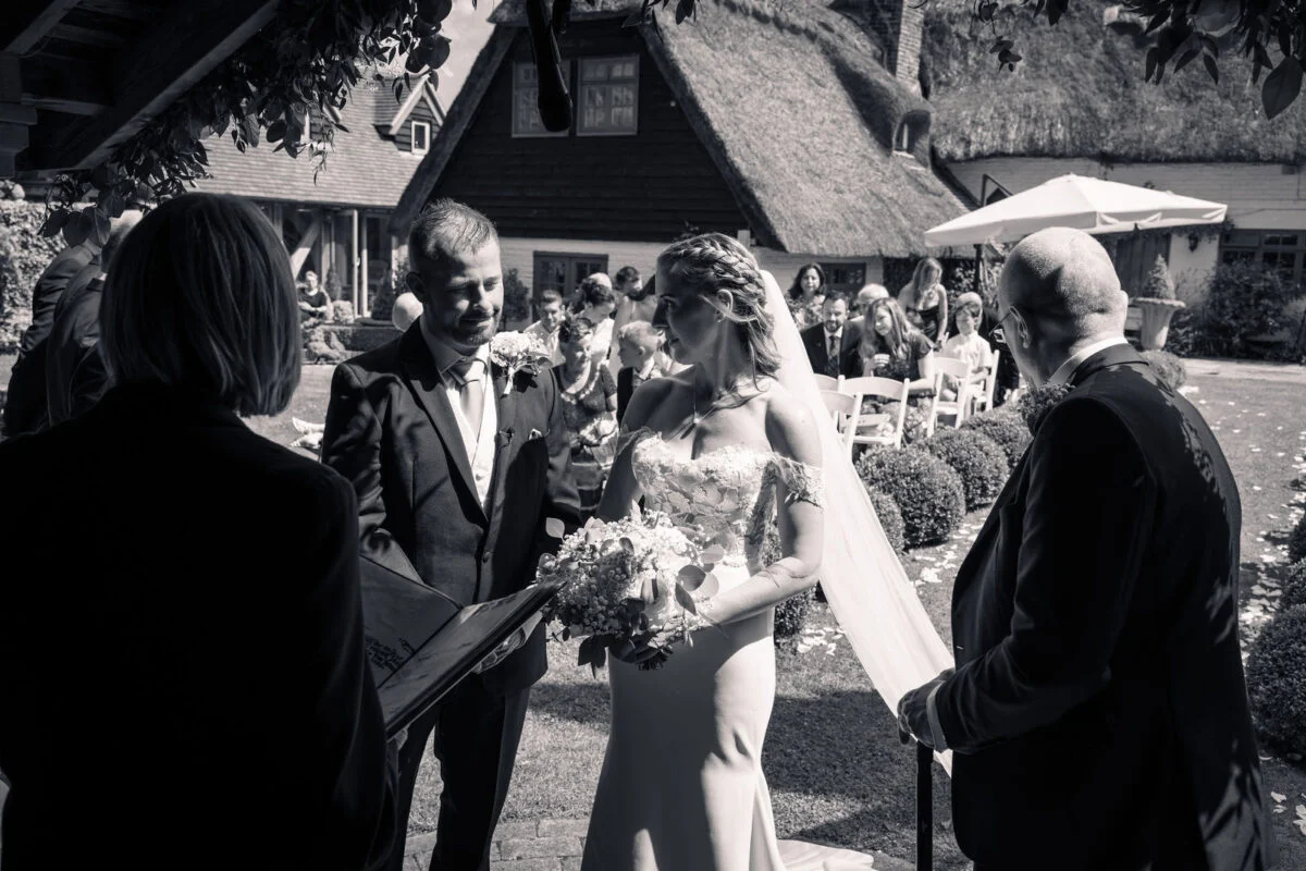 A bride and groom exchange vows during an outdoor ceremony on a sunny day, framed by seated guests and historic buildings.