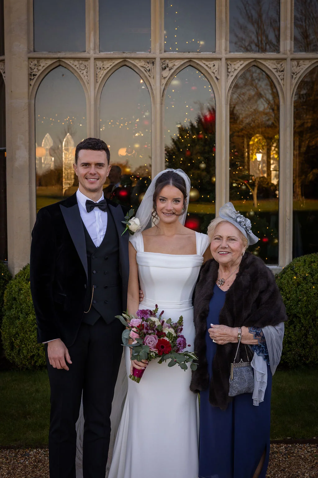 The bride and groom pose smiling outdoors with an elderly family member outside a stately manor house with large arched windows reflecting festive decorations.