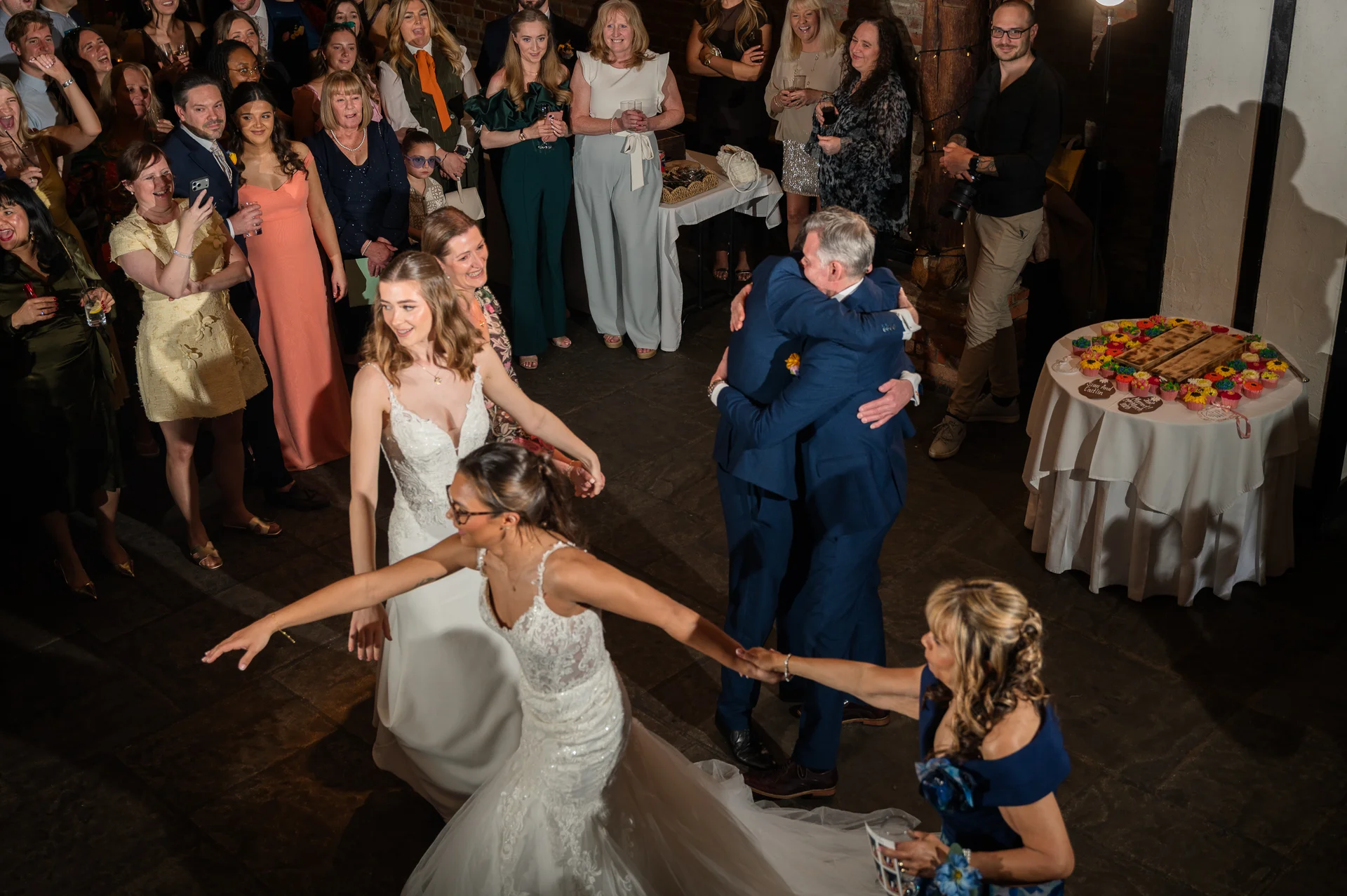 An elevated view captures two brides dancing together while guests look on, with two men embracing in the background near a dessert table.