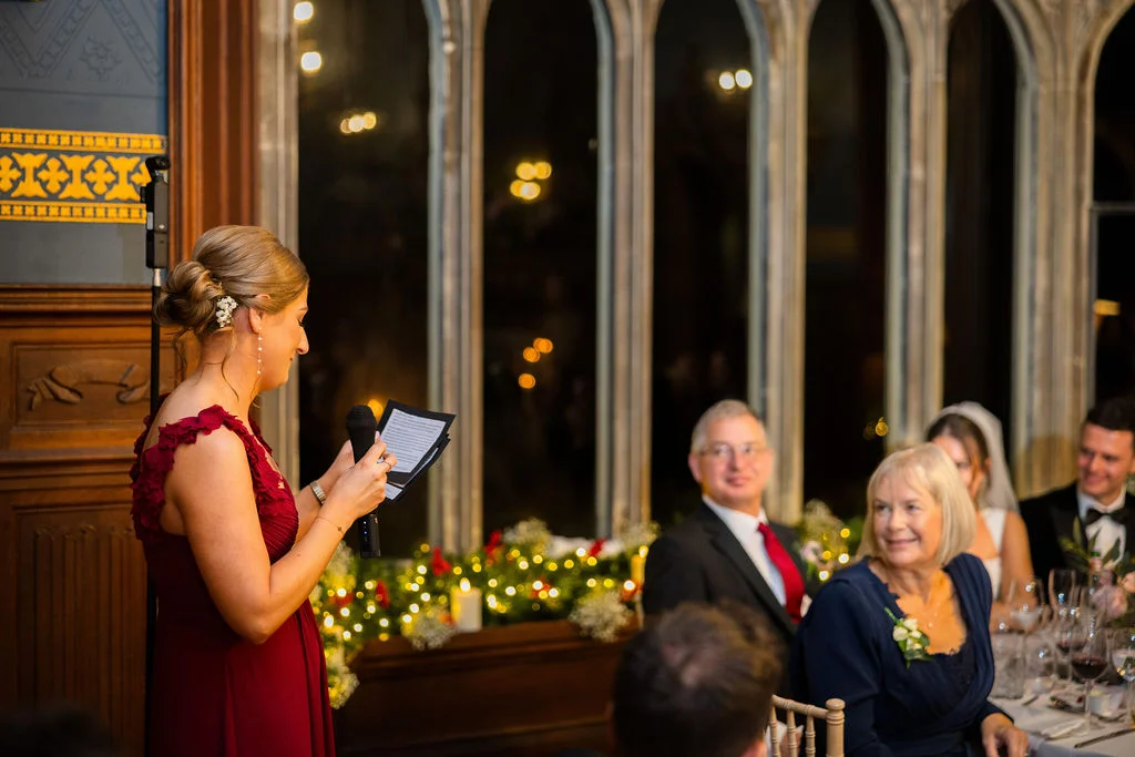 A bridesmaid in a burgundy dress delivers a speech holding a microphone and notes in front of tall arched windows and guests seated at tables.