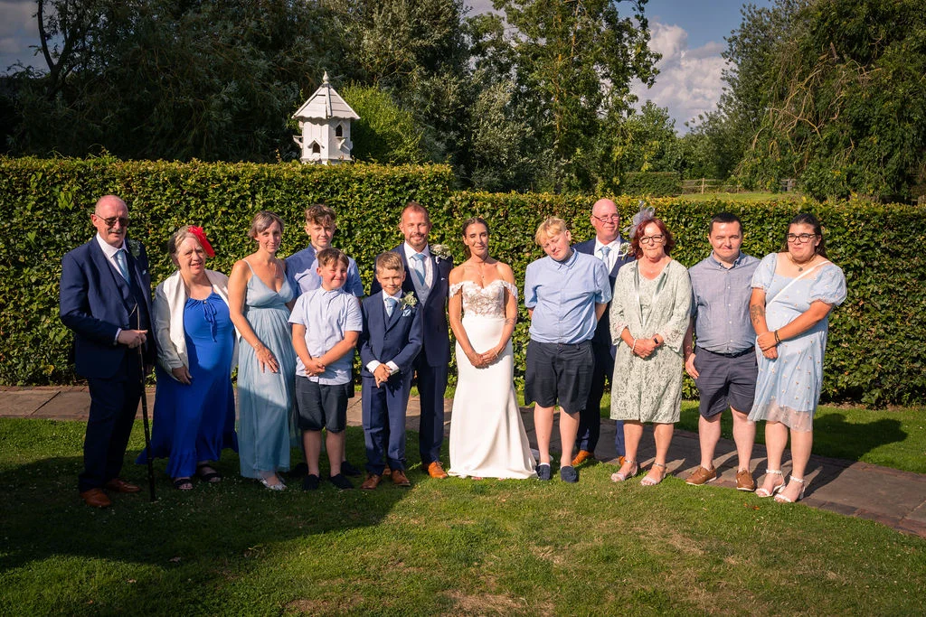 A large family group pose together outdoors on a sunny day in front of a tall green hedge at a wedding.
