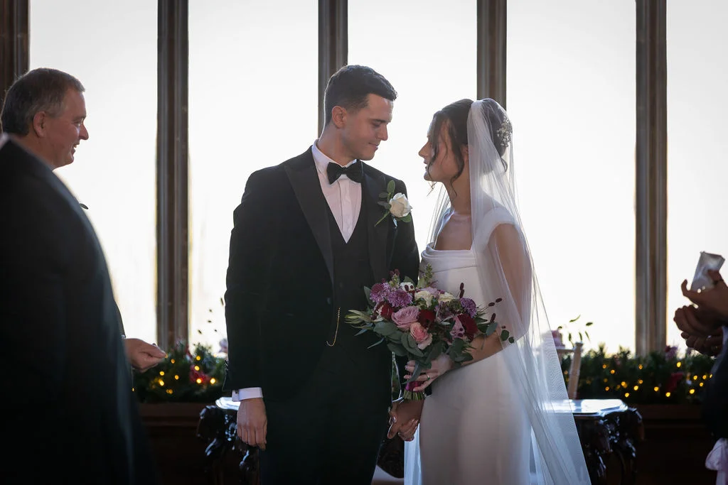 A newly married couple gaze lovingly at each other during their ceremony against tall windows, with the bride holding a rich burgundy bouquet.