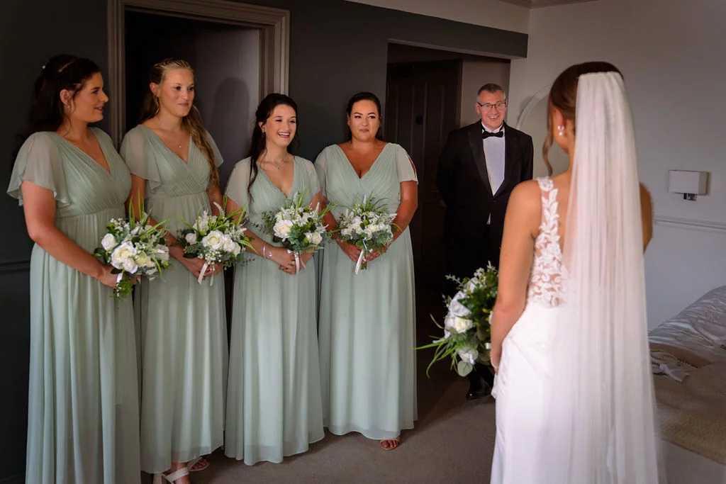 An emotional bride in a white lace gown and fingertip veil reveals her wedding look to her four bridesmaids in sage green chiffon dresses and a proud father in a tuxedo.