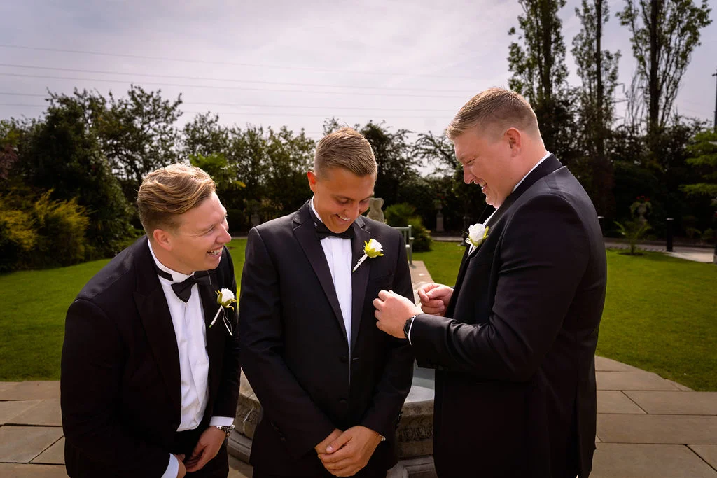 Three smiling groomsmen wearing black tuxedos and white rose buttonholes share a joyful moment outdoors on a paved patio.