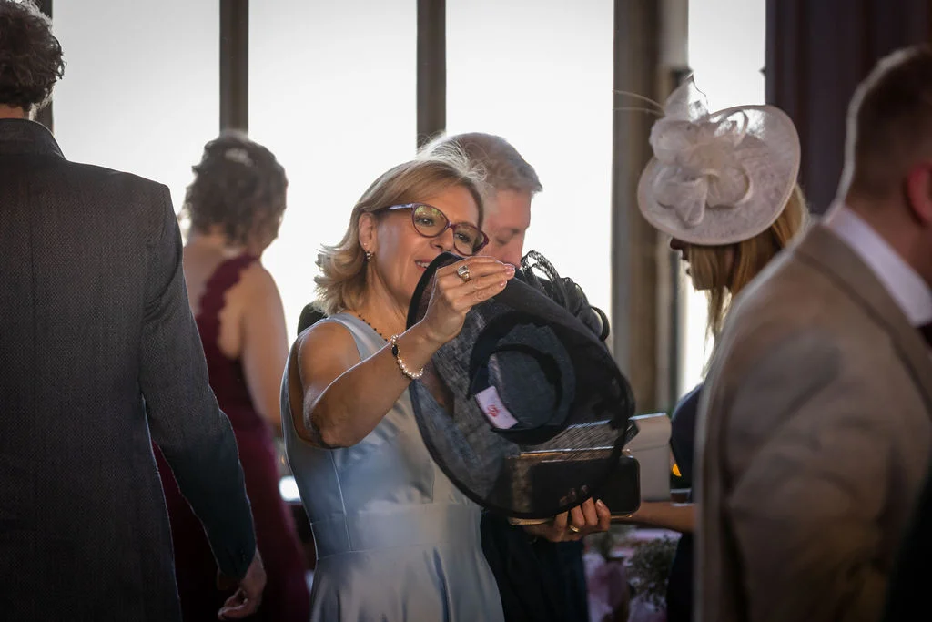 A smiling wedding guest in a light blue dress holds up and examines a large black fascinator amidst a gathering of mingling guests indoors.