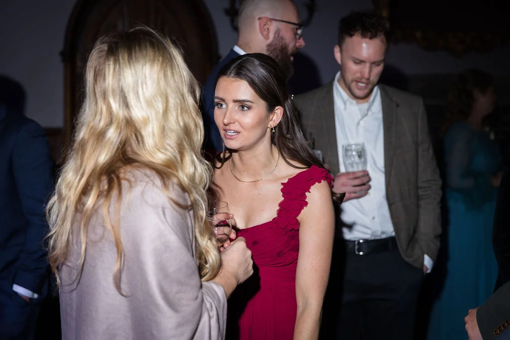 A guest in a burgundy dress chats with friends during an evening wedding reception as others mingle in the background.