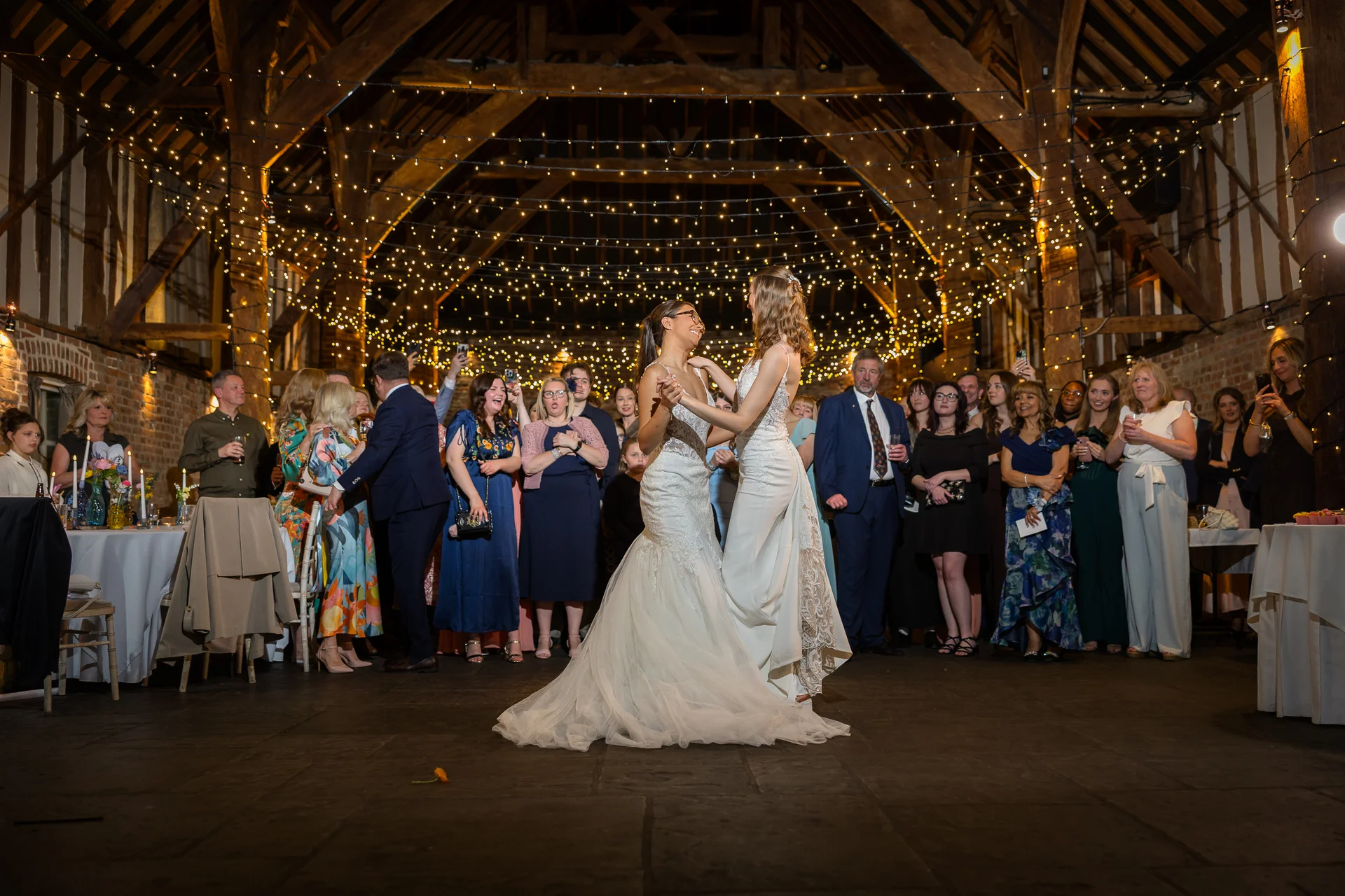 Two brides share their first dance in a rustic oak-framed barn illuminated by a stunning canopy of fairy lights while guests watch on.