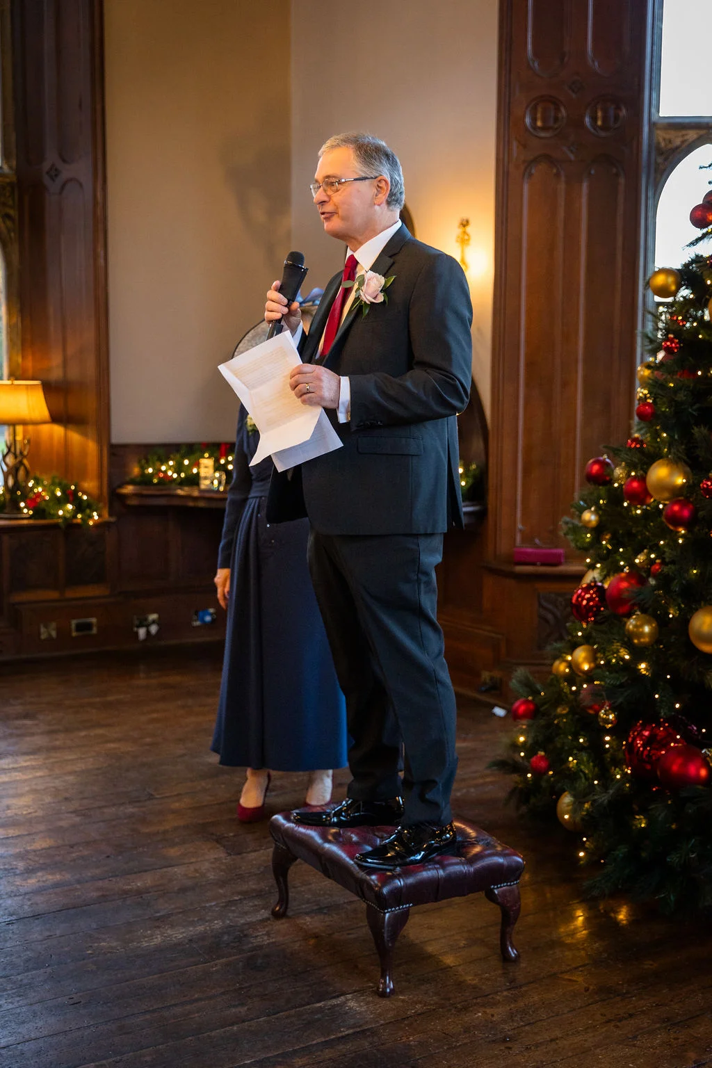 A man wearing a dark suit and red tie stands on a footstool while giving a speech at a winter wedding next to a decorated Christmas tree.