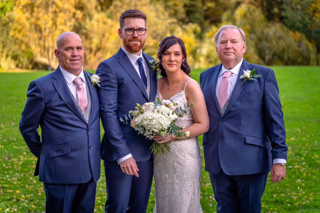 A formal outdoor wedding portrait features the bride holding a white bouquet alongside her groom and two older gentlemen in matching navy suits and pink ties on a manicured lawn.