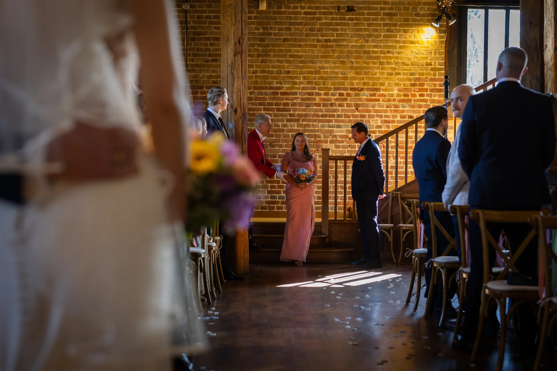 A bride in a pink dress walks down the aisle in a rustic barn wedding venue with exposed brick and wooden beams, watched by guests and groomsmen.