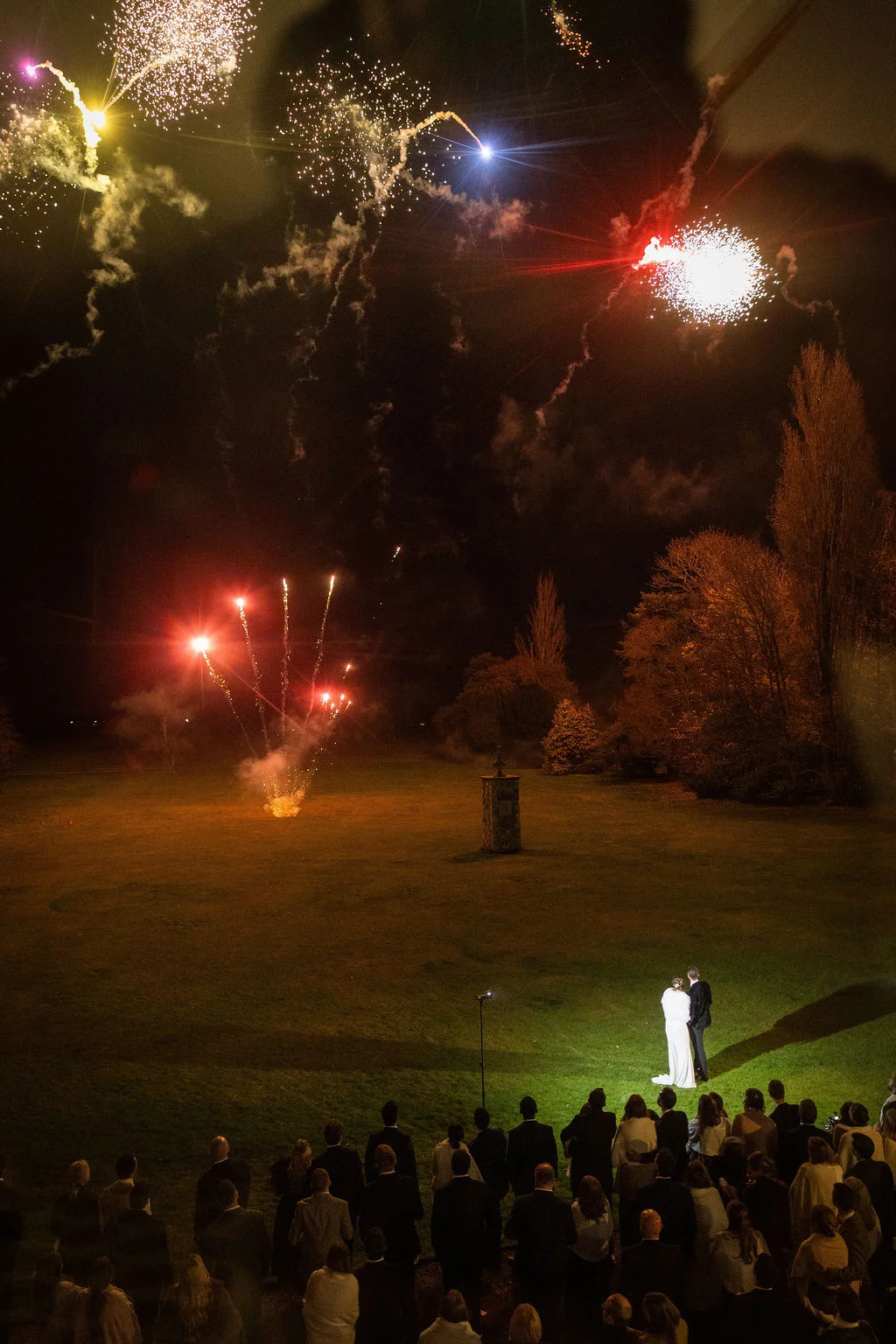 A newlywed couple stands on a sweeping lawn watching a dramatic nighttime fireworks display while surrounded by gathered wedding guests.
