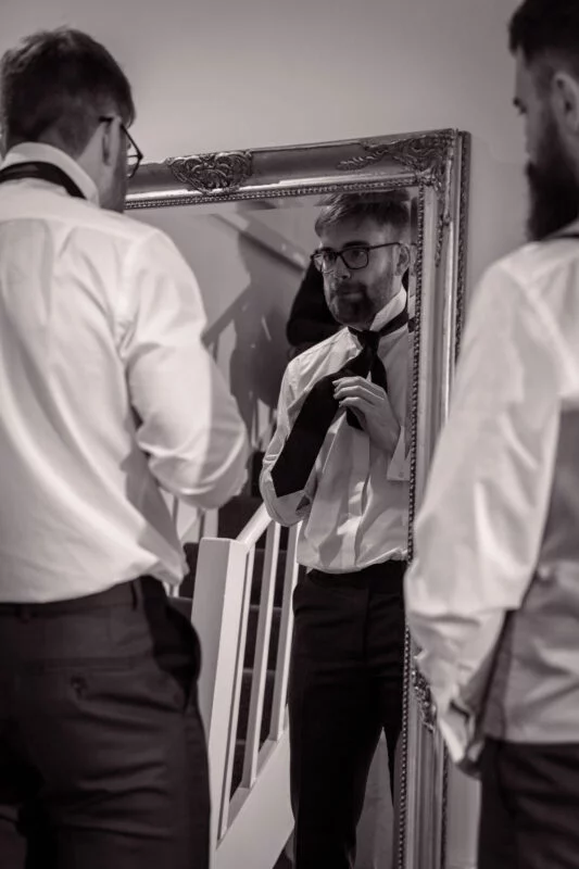 A groom standing in front of a ornate vintage mirror while adjusting his tie during morning preparations.