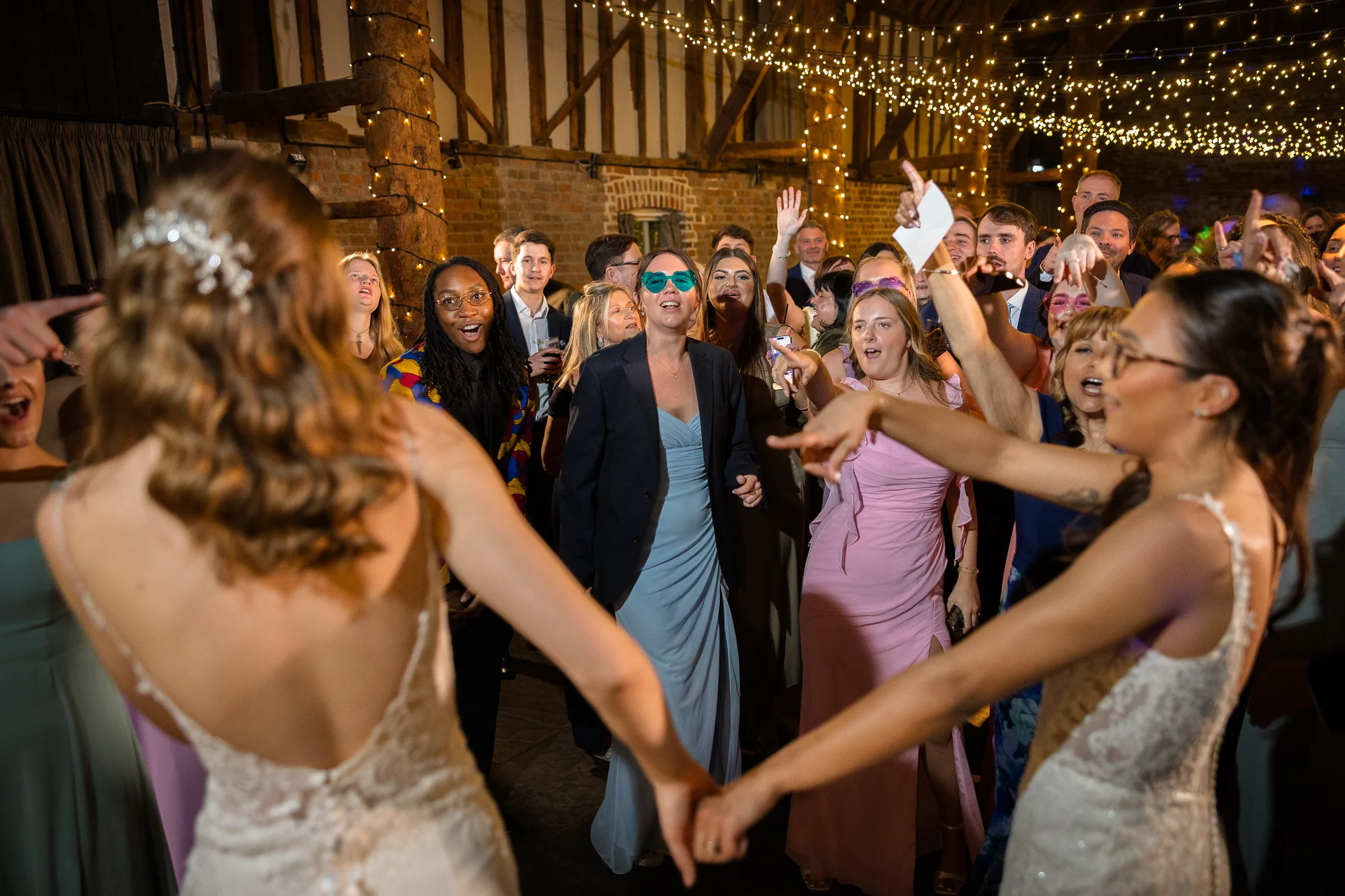 A joyful group of wedding guests cheer and dance in a rustic barn illuminated by a stunning fairy light canopy overhead.
