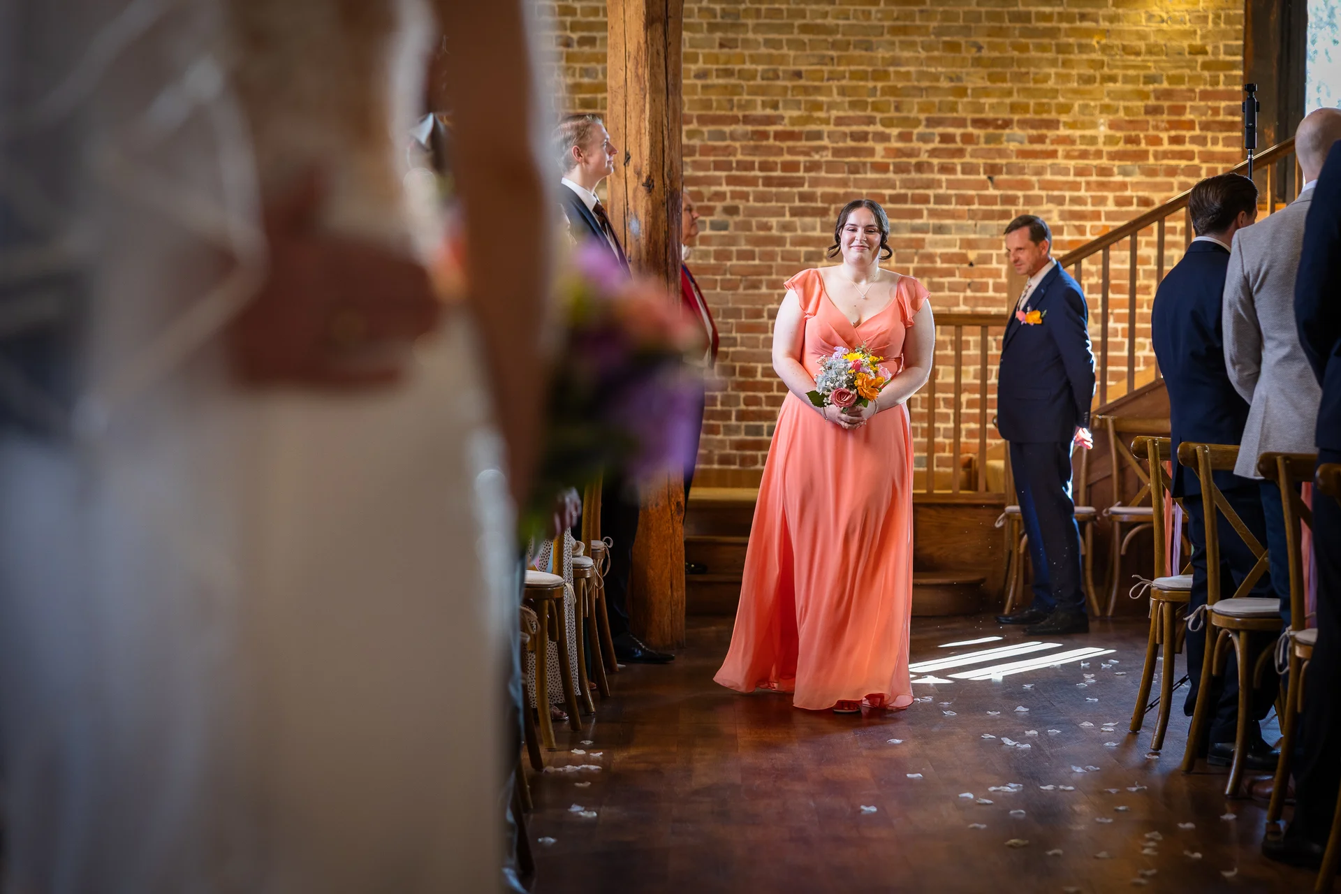 A smiling bridesmaid in a vibrant coral dress walks down the aisle inside a rustic barn venue with exposed brick walls.