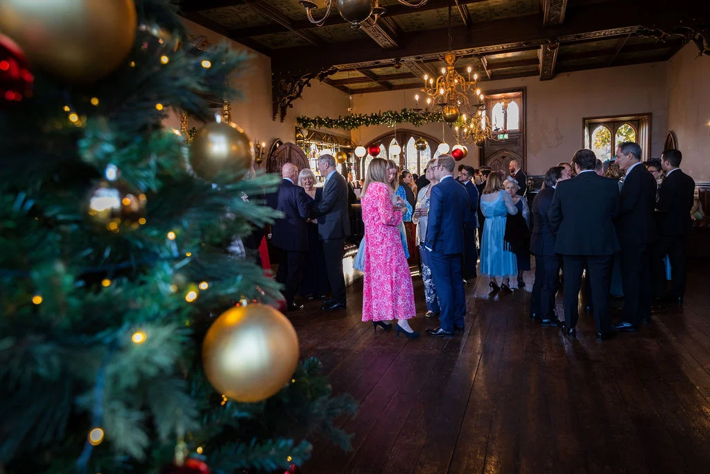 Wedding guests mingle during a festive drinks reception inside a stately home ballroom decorated with Christmas trees and warm lighting.