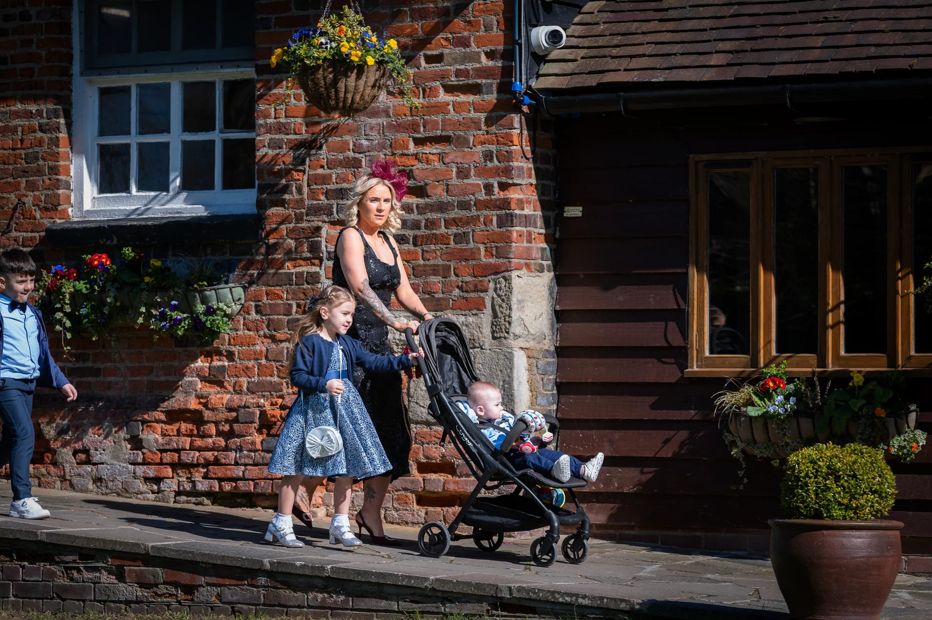Wedding guests including a woman wearing a fascinator and a young girl holding a small bag walk past a traditional red brick building while pushing a baby in a stroller.