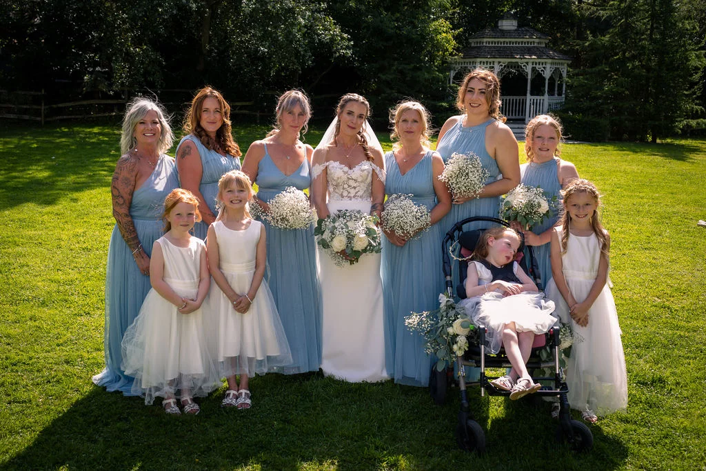 A bride in a lace-trimmed gown poses outdoors on a sunlit lawn surrounded by her bridesmaids in dusty blue dresses and young flower girls.