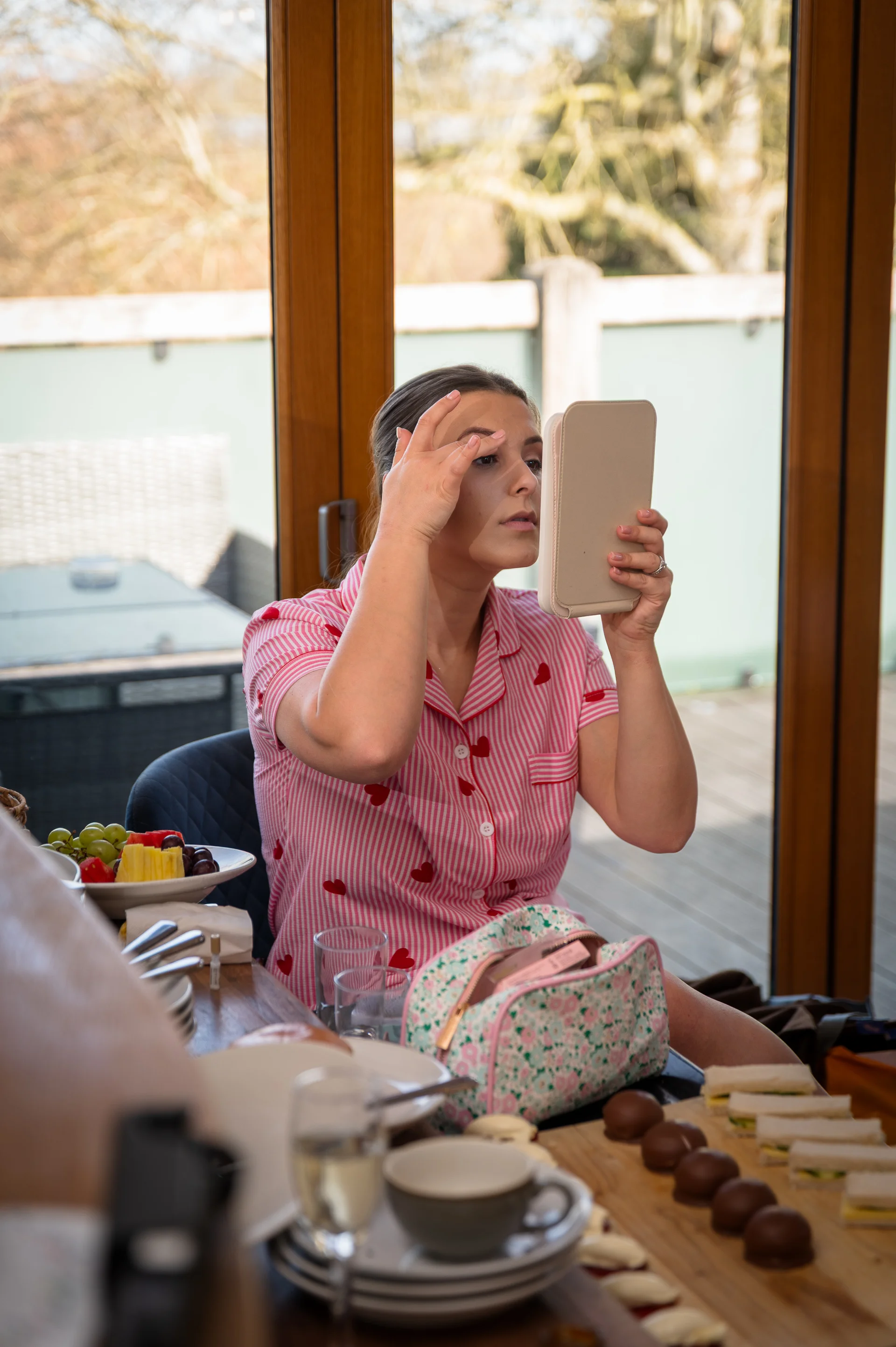 A bride sits at a table near a large glass door in her morning pyjamas, checking her makeup in a small handheld mirror during wedding preparations.