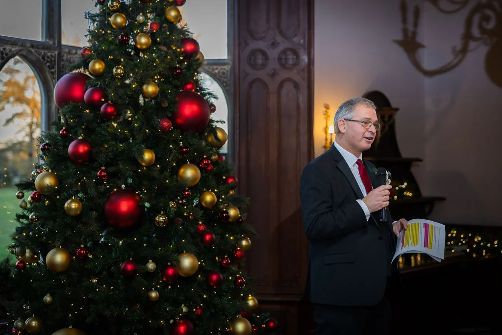 A mature man in a suit delivers a speech holding notes and a microphone next to a large Christmas tree decorated with red and gold baubles.