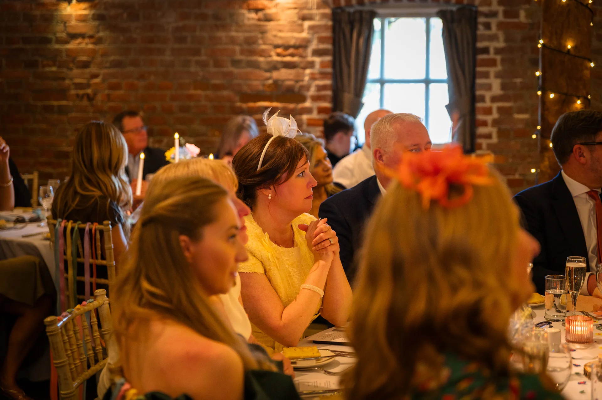 Guests attentively listen during the wedding speeches inside a rustic venue with exposed brick walls and warm candlelight.