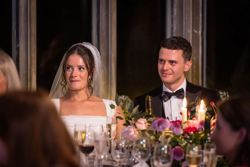 A newlywed bride and groom sit together at the top table during the wedding breakfast, smiling and listening to speeches by candlelight.