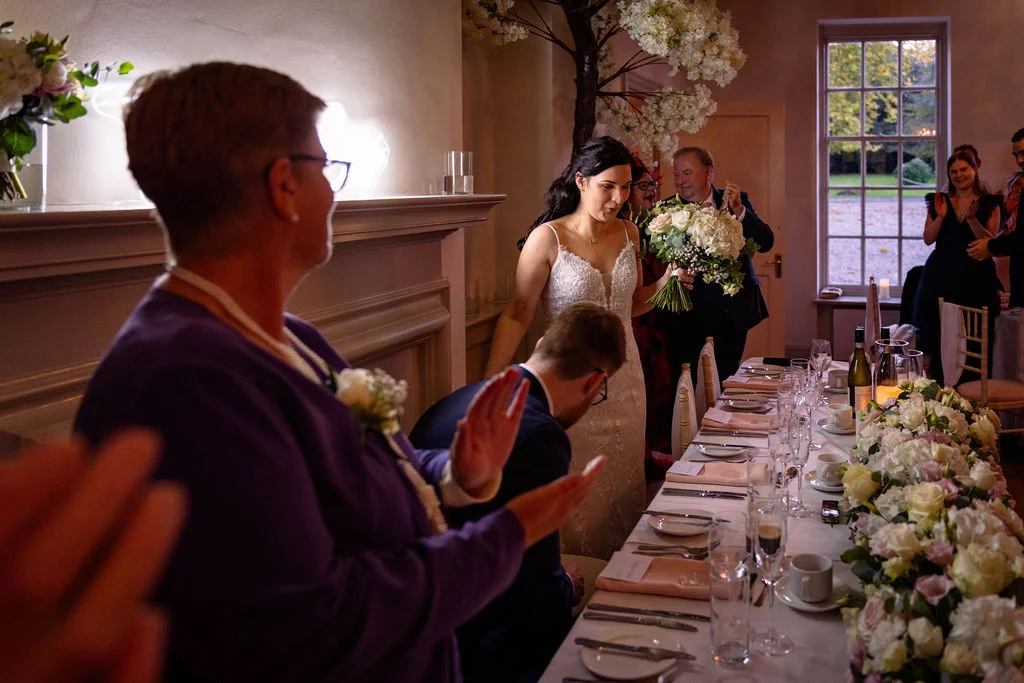 A smiling bride holding a floral bouquet enters the wedding reception room while seated guests applaud at a beautifully decorated long banquet table.