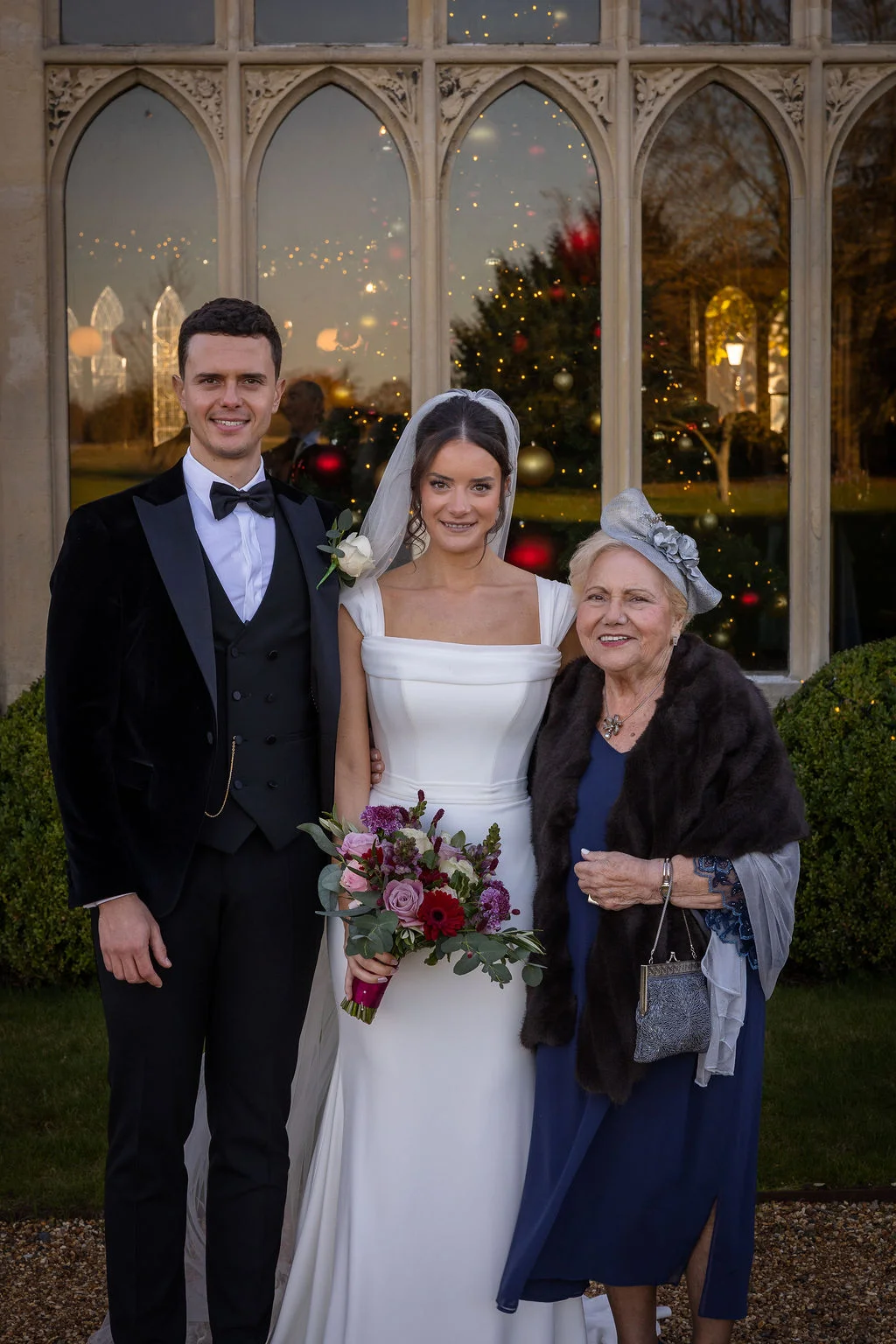 A newlywed couple poses outside a stately stone venue with tall arched windows for a family portrait alongside an elderly family member during a winter wedding.