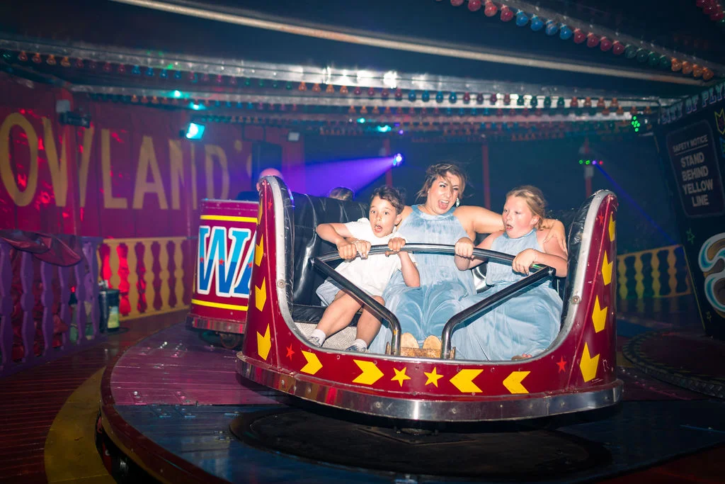 Wedding guests laughing and screaming with excitement as they ride a brightly lit funfair attraction at an evening reception.