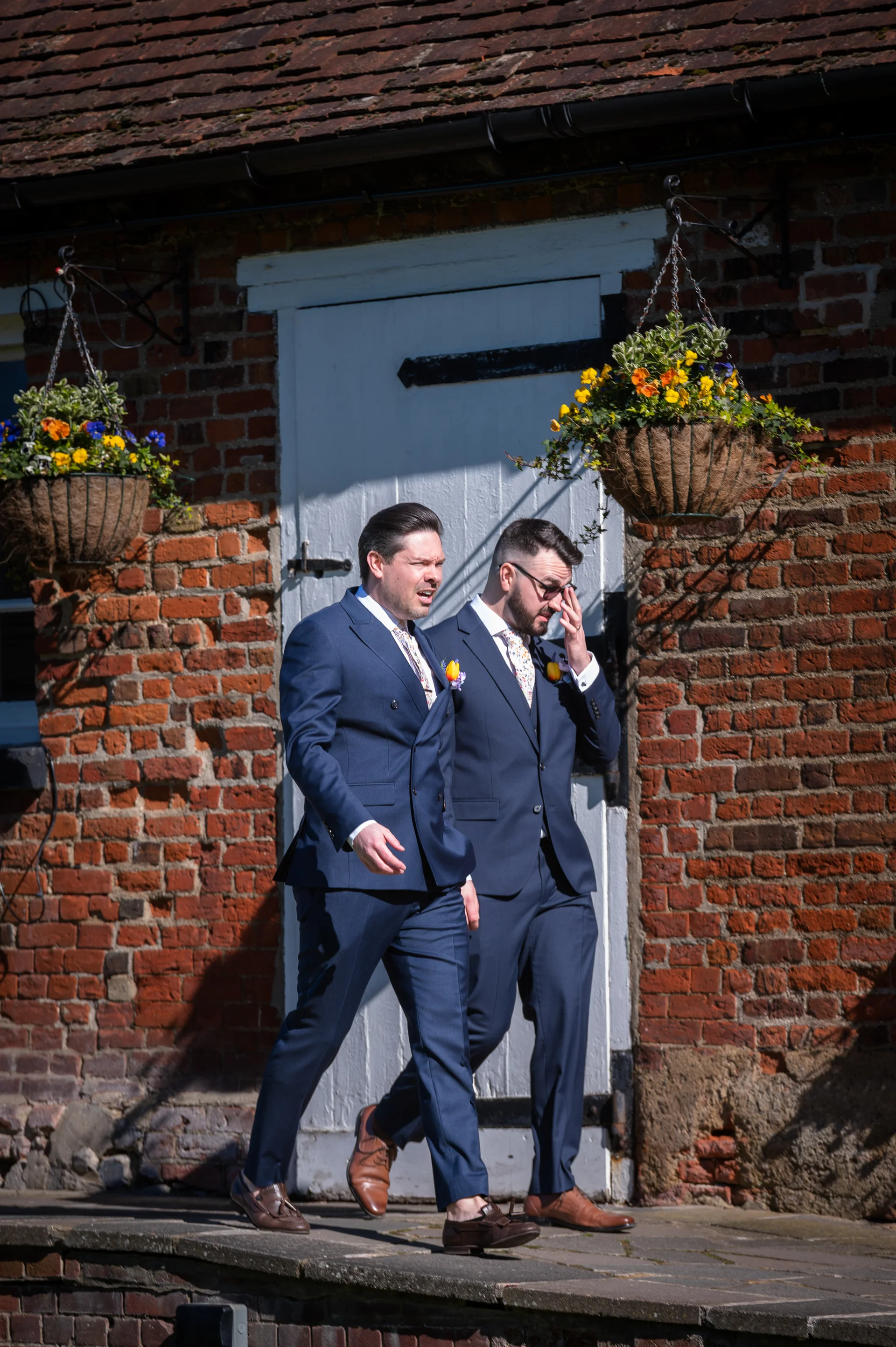 Two grooms in sharp navy suits walk together outside a rustic red brick building on a bright sunny wedding day.