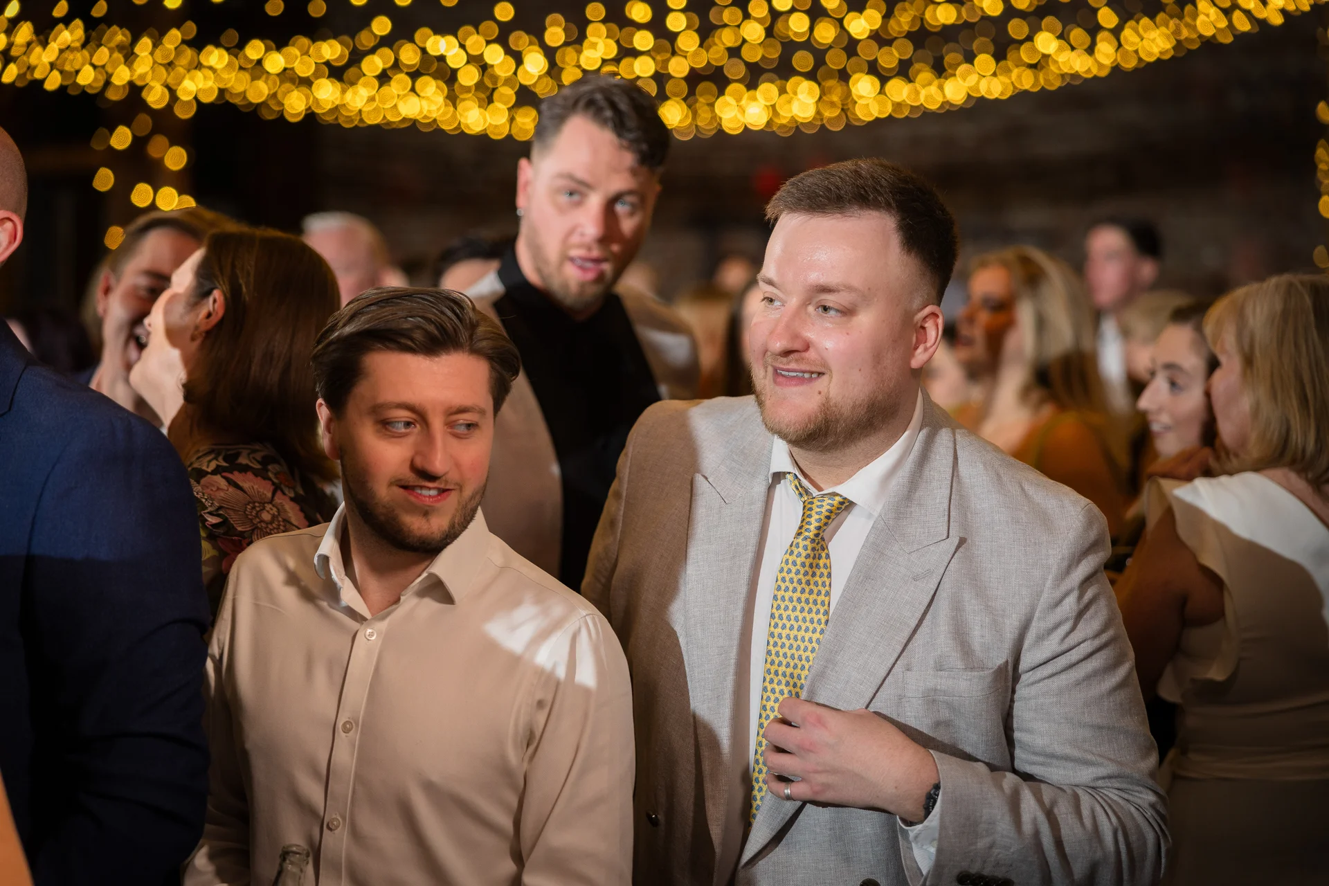 A candid shot of wedding guests mingling at an evening reception under a warm canopy of string lights.