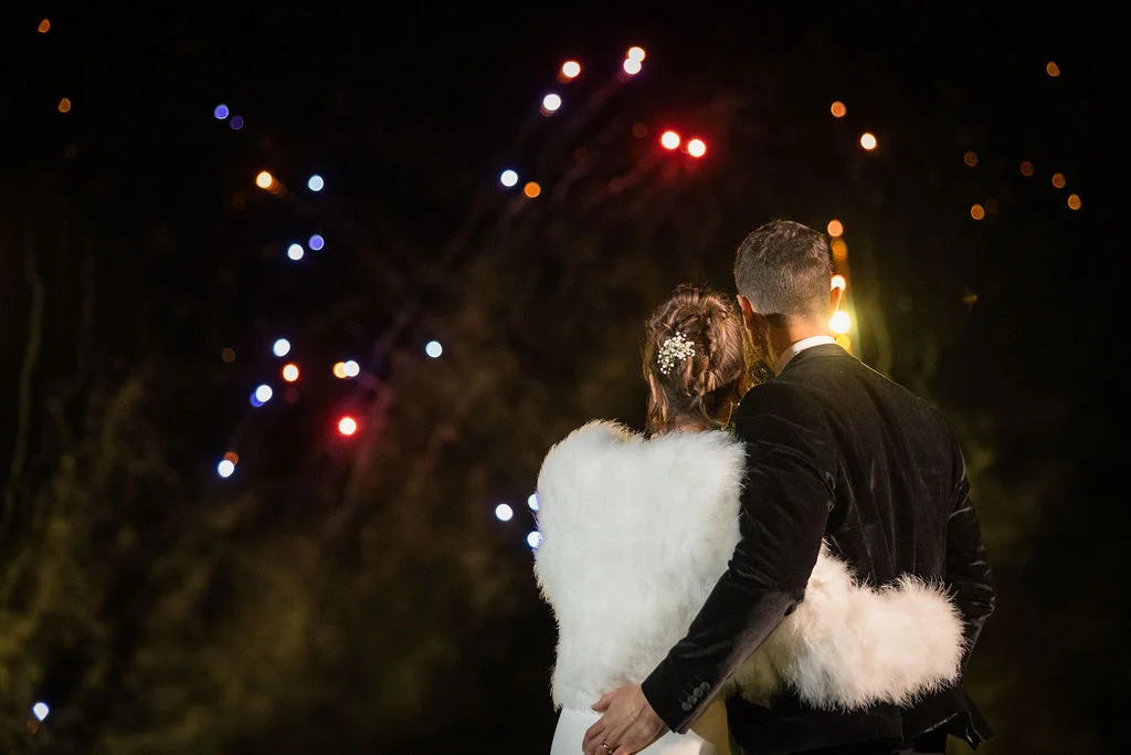 A newlywed couple viewed from behind embraces while watching a vibrant fireworks display light up the night sky.