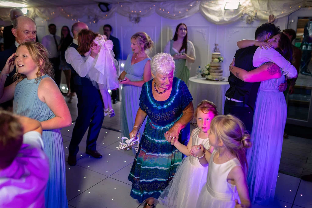 Guests and family members joyfully dance together on an illuminated dance floor inside a marquee reception lit with purple mood lighting.