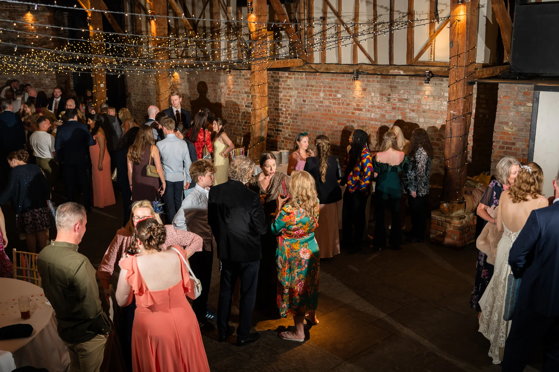 Guests mingle and chat during an evening reception inside a rustic brick barn illuminated by a canopy of twinkling string lights.
