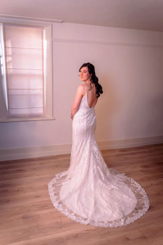 A stunning bride in a lace a-line wedding dress with spaghetti straps and a long train poses gracefully indoors, glancing back toward the camera near a bright window.