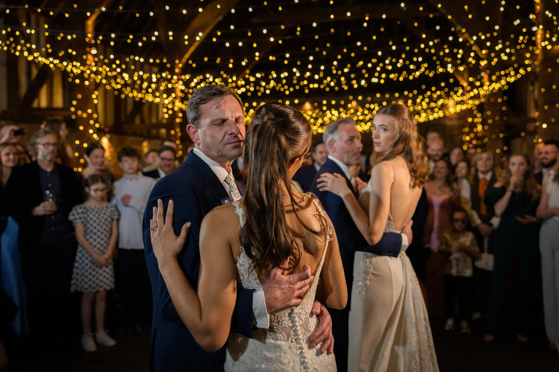A father and bride share an emotional embrace during their first dance under a magnificent canopy of twinkling fairy lights inside a rustic oak-framed barn.
