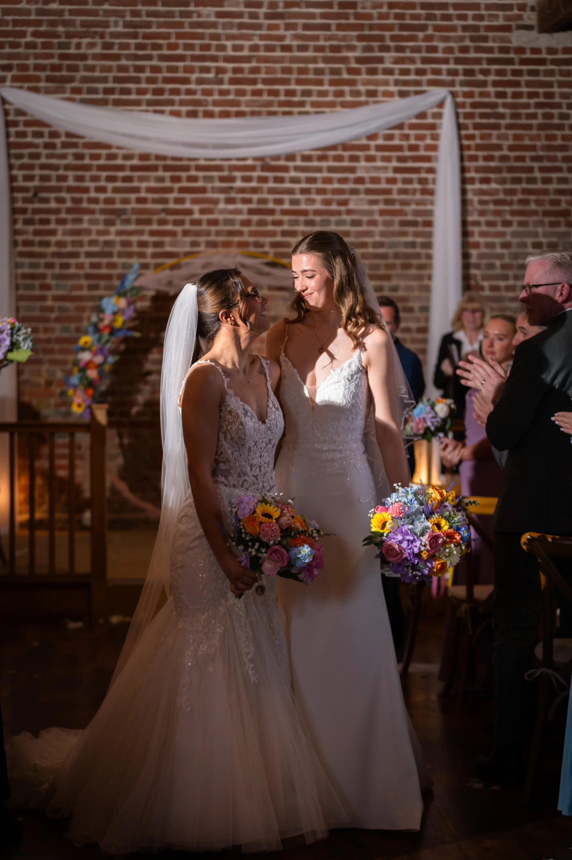 Two brides in stunning white wedding dresses lovingly gaze at each other while holding colorful floral bouquets inside a rustic brick barn venue.