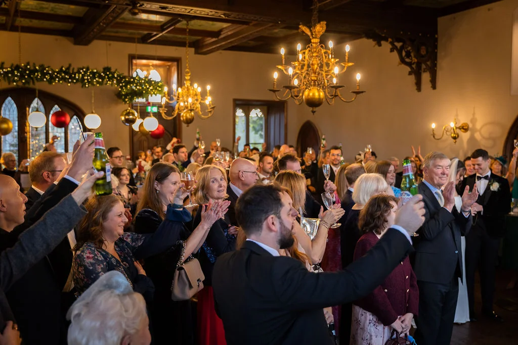 A large wedding crowd raises glasses and bottles in a grand, chandelier-lit hall during the reception speeches.
