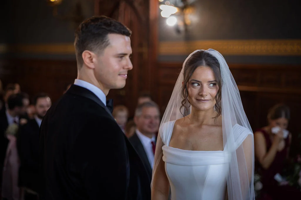 A newly married couple share a tender glance during their traditional indoor wedding ceremony, with the bride wearing an elegant white gown and veil.