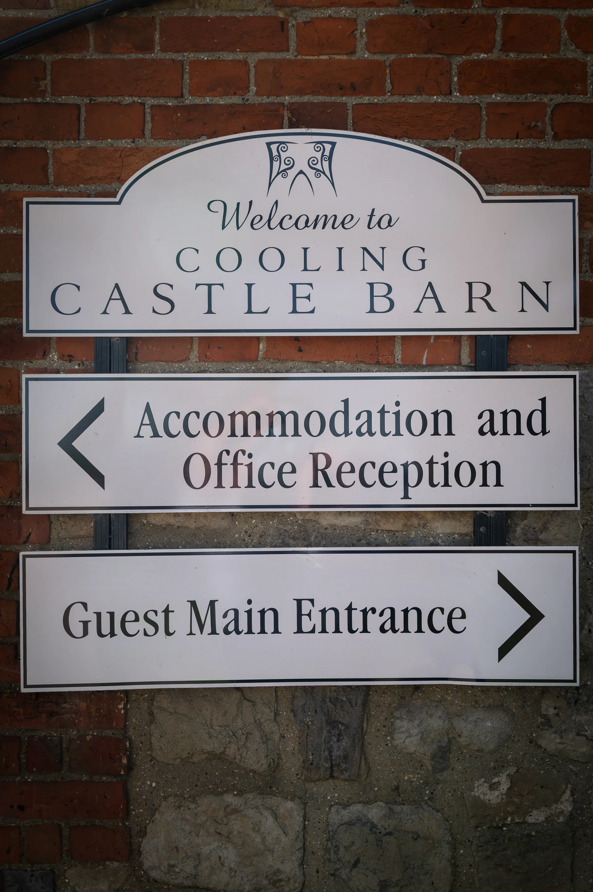 A clear directional sign welcoming guests to Cooling Castle Barn, mounted against a textured red brick and stone wall.