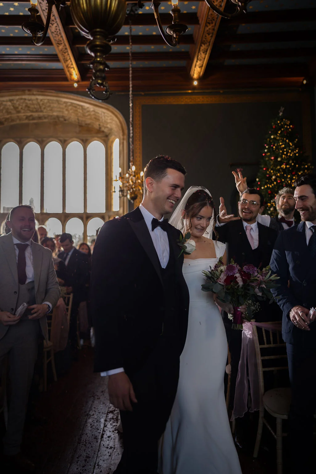 A newly married couple smiling happily as they walk up the aisle surrounded by cheering guests during a confetti exit inside a historic venue with high vaulted ceilings.