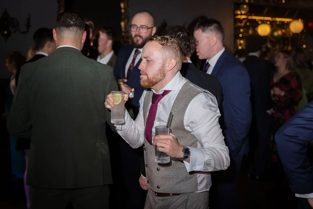 A groom or groomsman in a tweed waistcoat and patterned tie holds a highball glass during a lively wedding drinks reception.