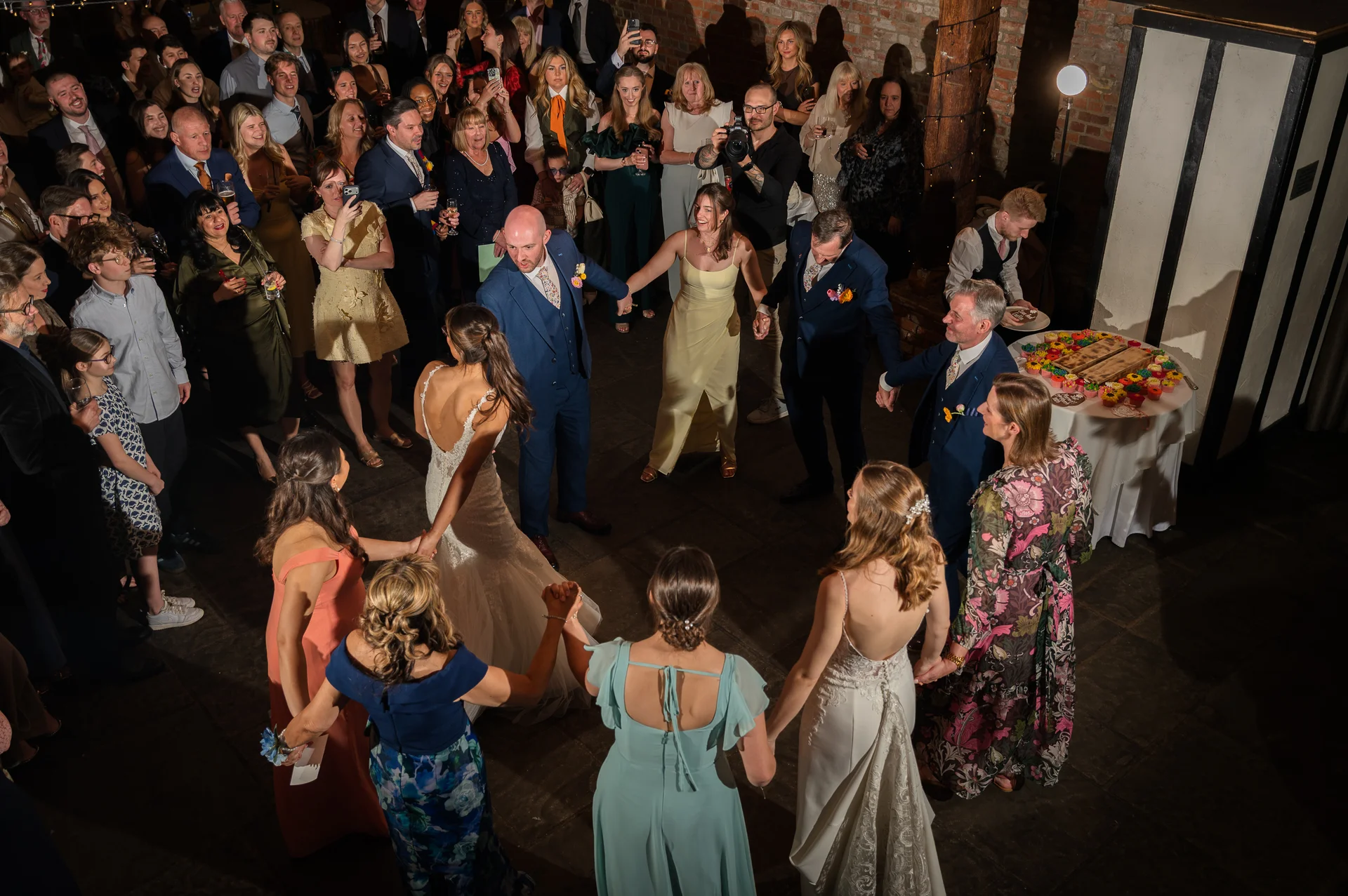 An elevated wide shot captures a lively circle of guests and the newlyweds holding hands and dancing together during an evening wedding reception inside a rustic brick barn venue.