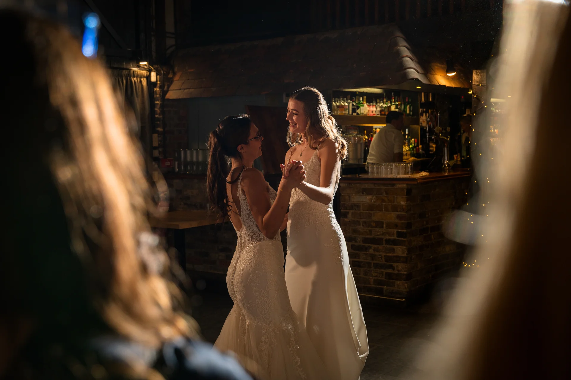 Two brides share a joyful first dance together in a dimly lit pub reception venue framed by guests in the foreground.