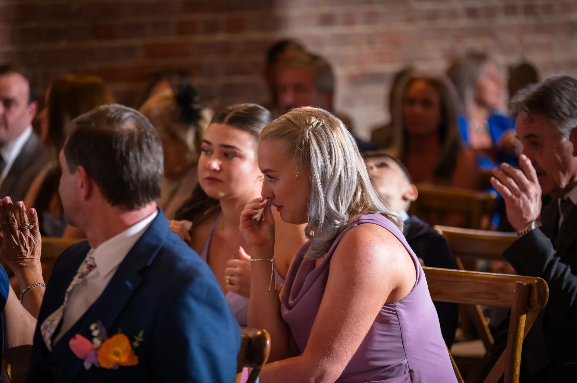 A guest wiping a tear of joy from her eye during an emotional wedding ceremony set against an exposed brick wall.