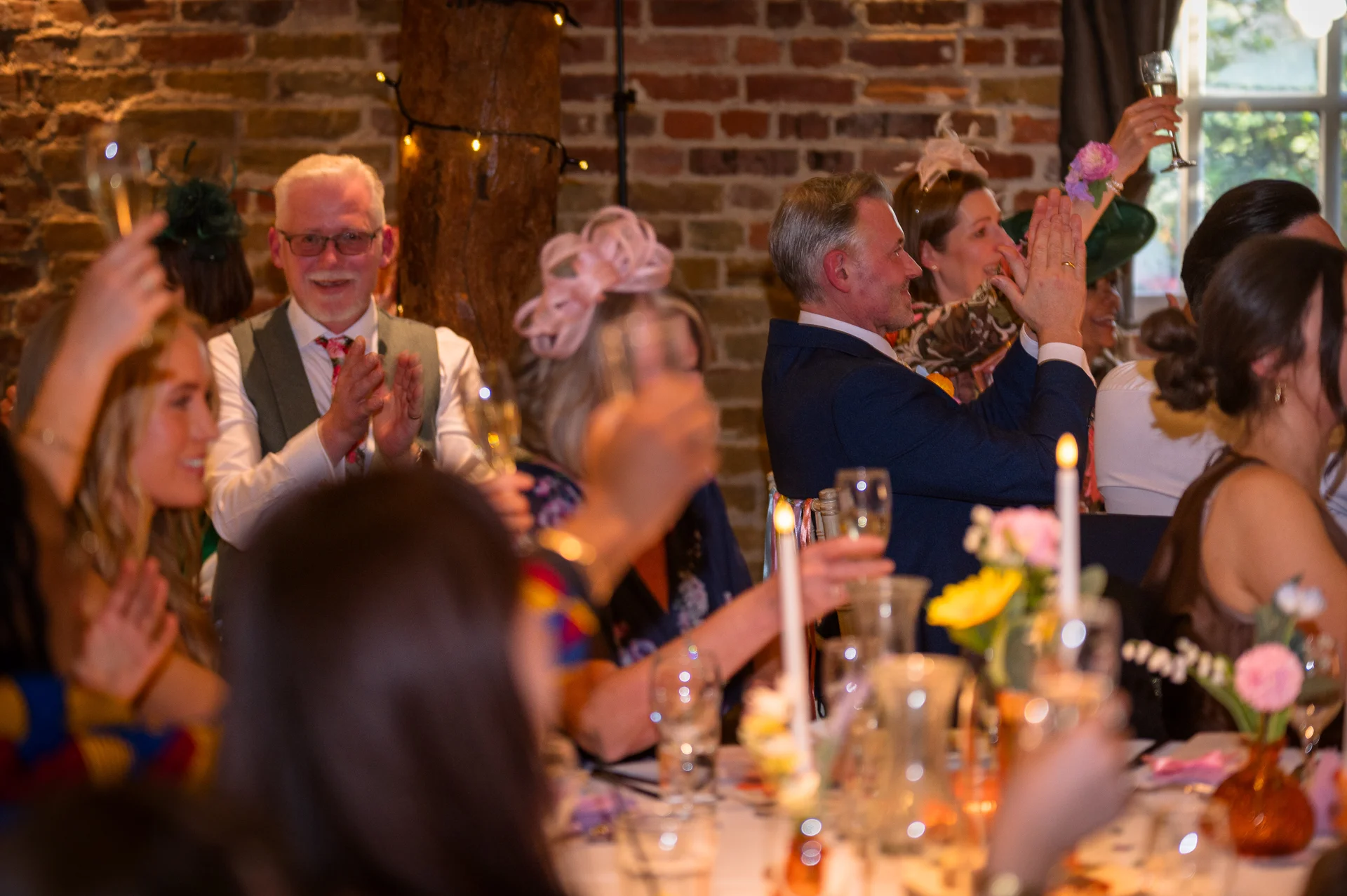 Wedding guests enjoy the wedding breakfast and laugh during the speeches in a rustic barn venue with exposed brick walls.