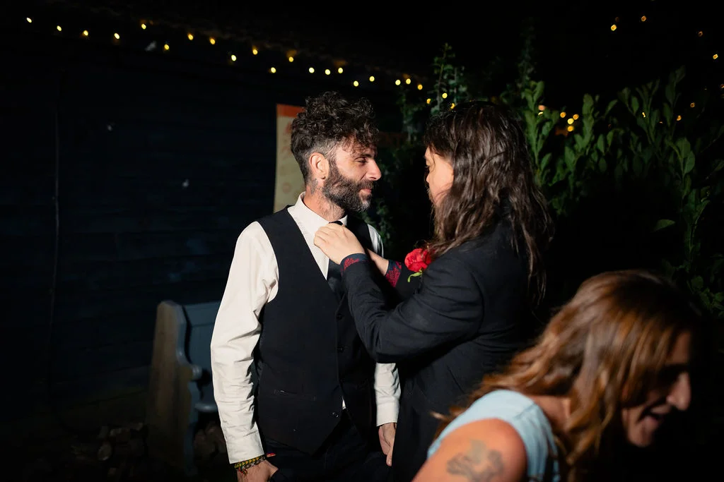 One groom gently adjusts the tie of another groom during an evening celebration lit by overhead fairy lights.