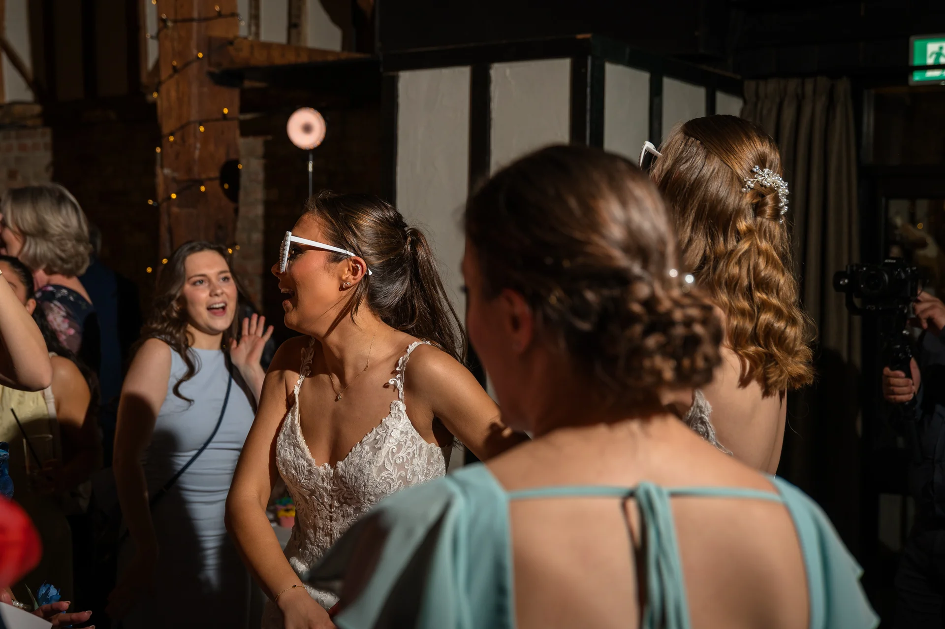 A laughing bride wearing fun white sunglasses and a lace gown celebrates on the dance floor surrounded by wedding guests during an evening reception.