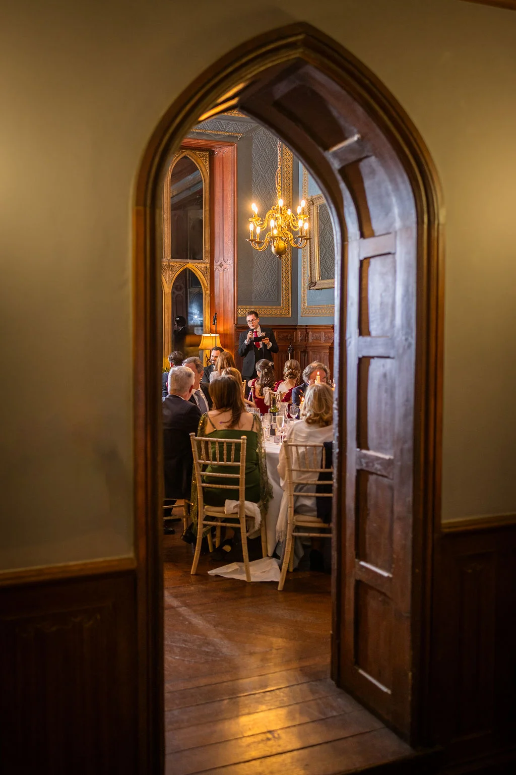 Guests are seated around tables enjoying wedding speeches in a wood-panelled room, viewed through a traditional stone and wood archway.